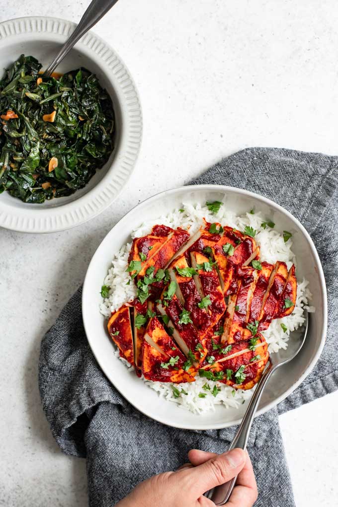 a bowl of rice topped with sliced barbecue tofu with a side of smoky collard greens, a hand is reaching into the tofu with a fork