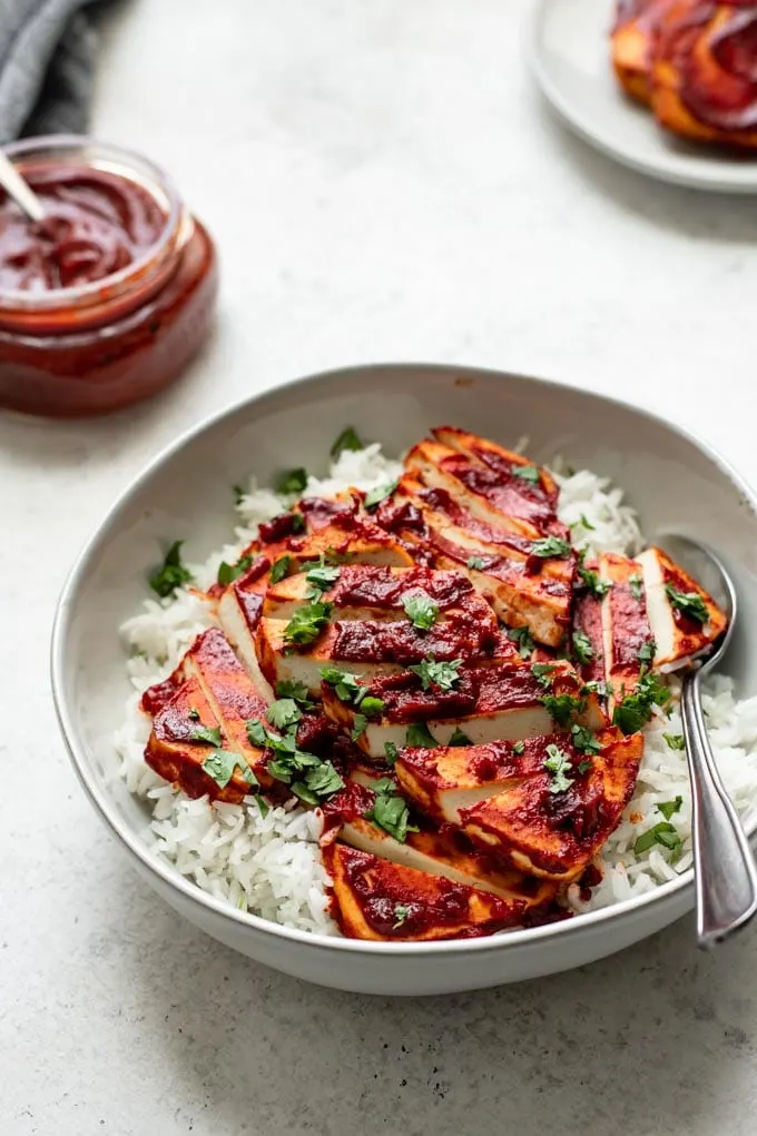 side view of a bowl of barbecue tofu over rice, garnished with cilantro. A jar of barbecue sauce is in the background as well as a few slices of barbecue baked tofu on a plate
