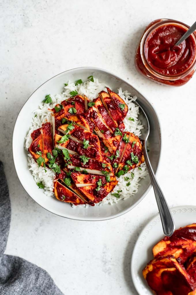 barbecue baked tofu in a bowl over rice, with a plate with extra BBQ tofu and some barbecue sauce