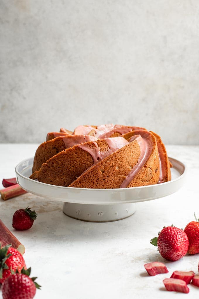 almond rhubarb bundt cake with a strawberry rhubarb glaze on a stand with strawberries and rhubarb surrounding it