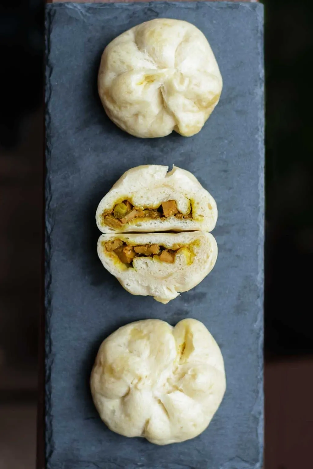 3 curry seitan siopao on a black slate with the middle one cut open. Dark background.