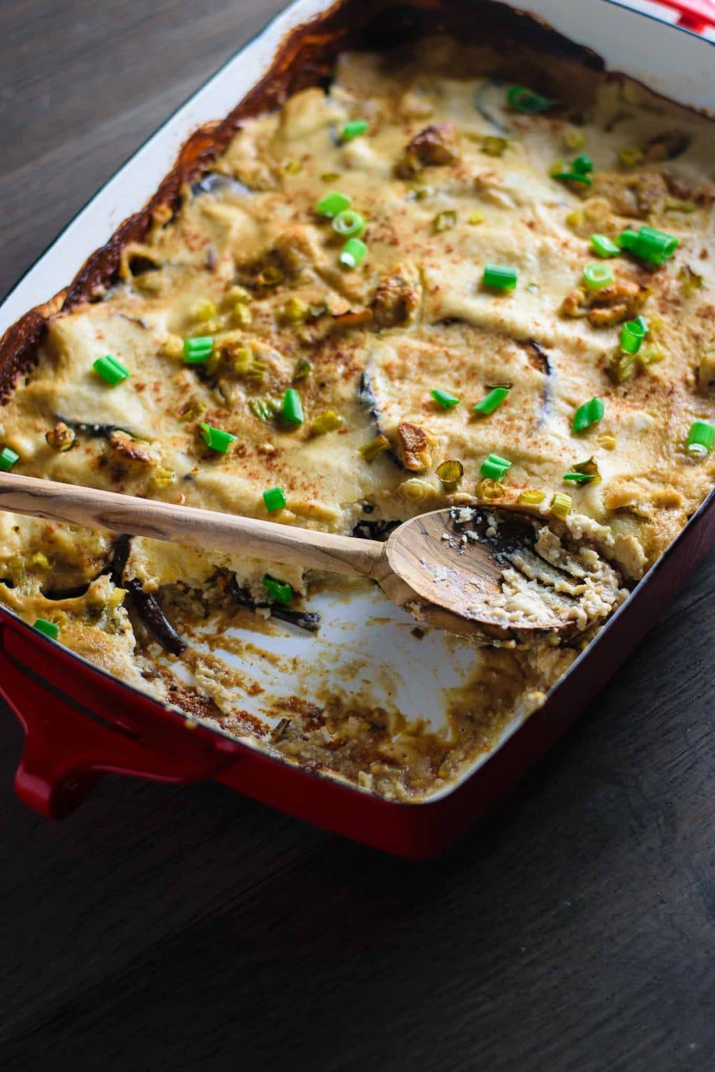 a wooden spoon in the vegan cheesy eggplant casserole dish, with a serving taken out, shot from an angle and lit from behind