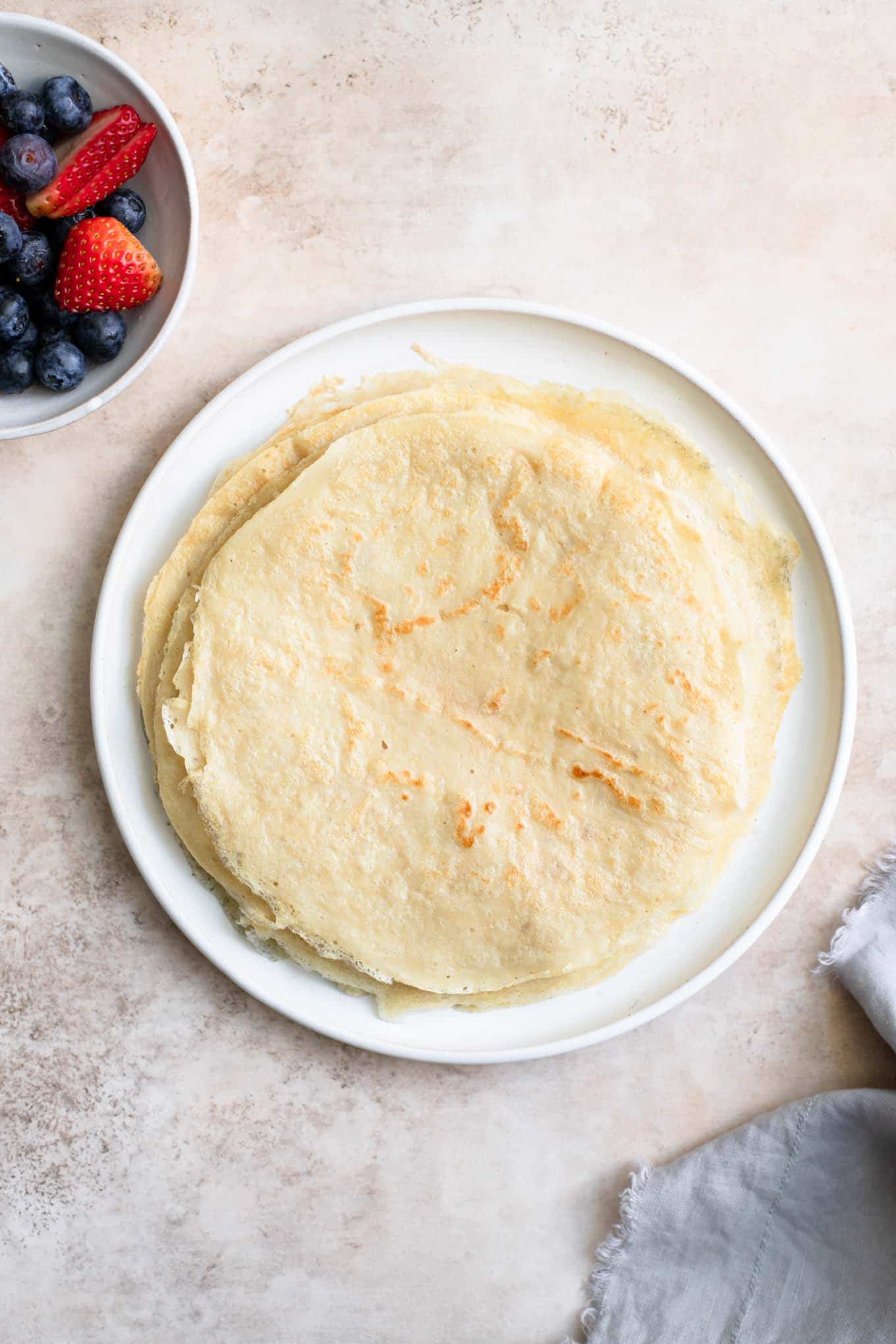 stack of crepes on a plate with a small bowl of fruit on the side