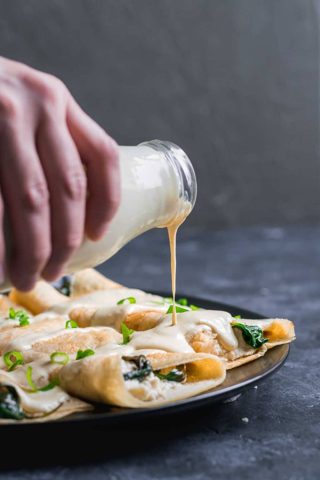 pouring vegan hollandaise sauce over savory crepes filled with almond cheese and sauteed spinach. Garnished with scallions