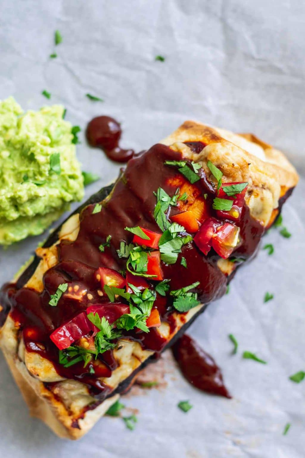 Vegan smothered burritos with red sauce, topped with extra red chile sauce after baking, diced cherry tomatoes and cilantro, and served with mashed avocado