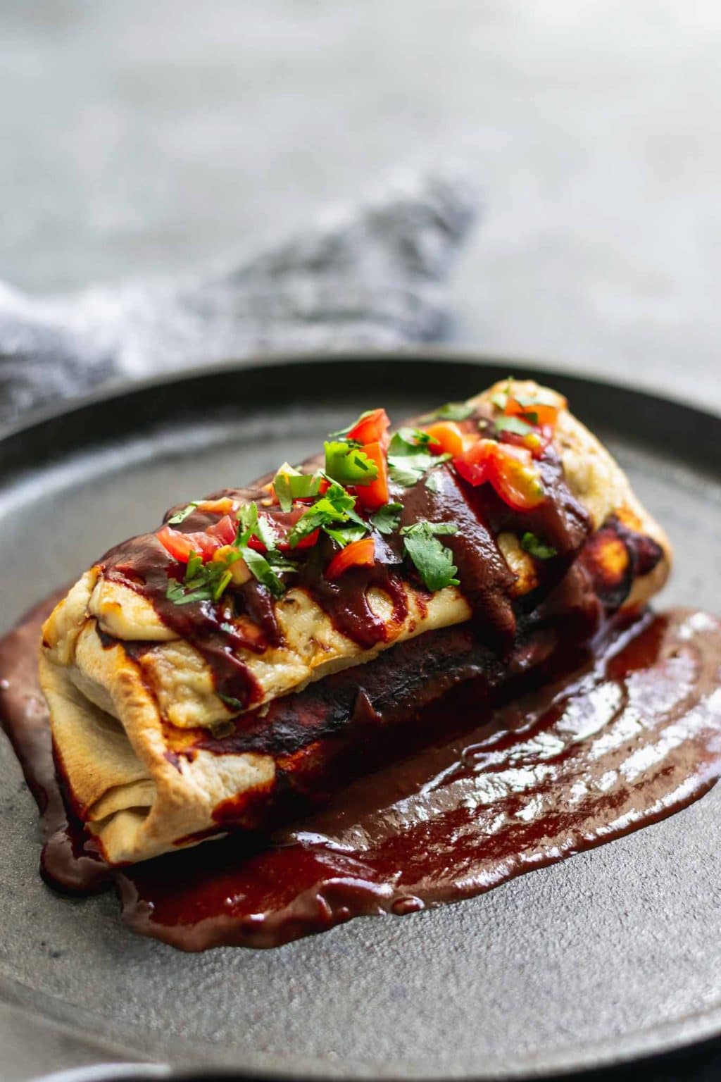 Vegan smothered burritos with red sauce topped with melted cheese and topped with a lot of cherry tomatoes and cilantro on a bed of red sauce.