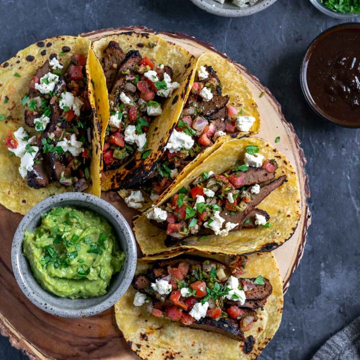 Vegan seitan carne asada tacos topped with pico de gallo and vegan queso fresco with guacamole and extra marinade, queso fresco, and cilantro to serve. 5 tacos assembled on a platter.