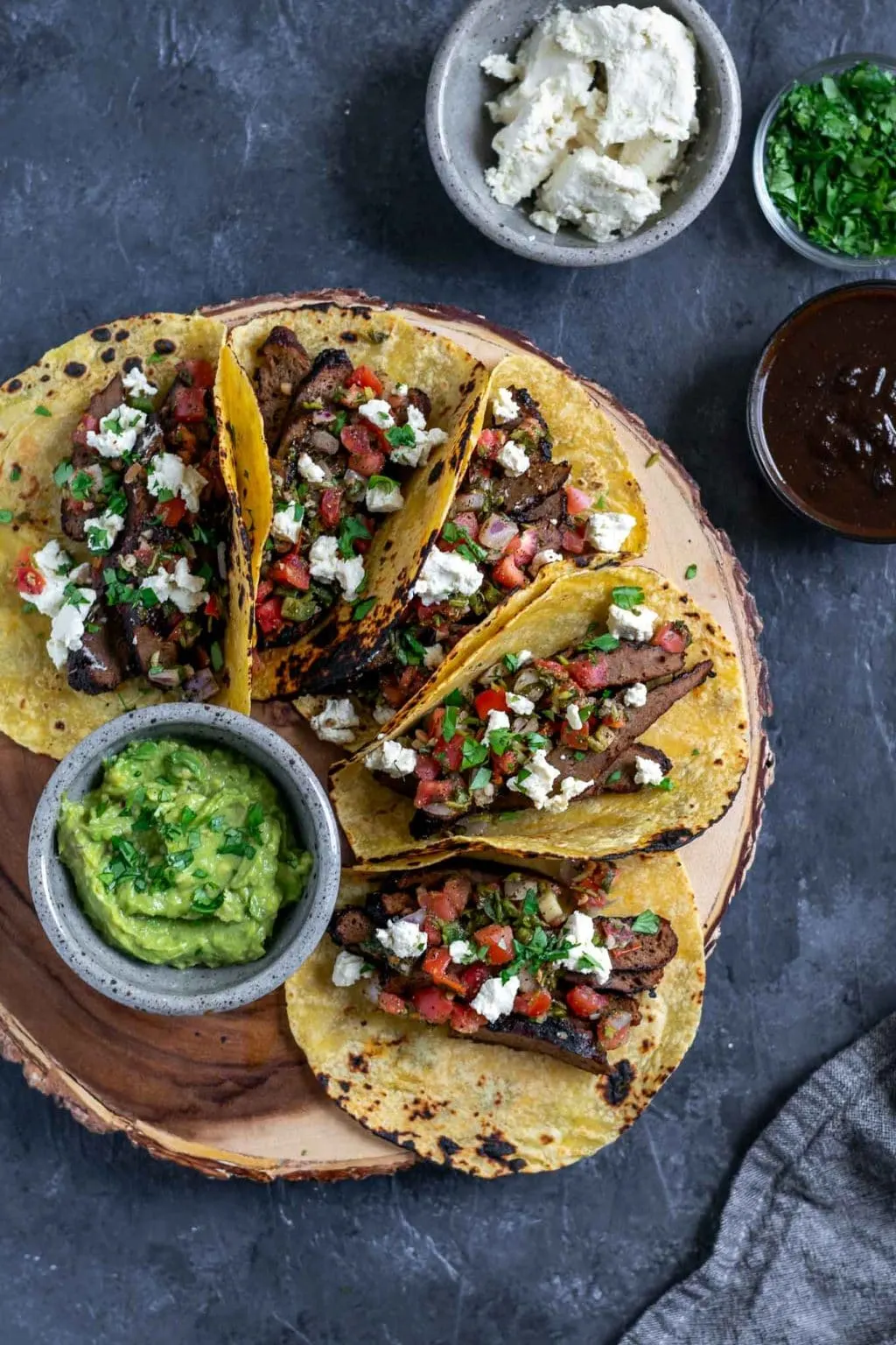 Vegan seitan carne asada tacos topped with pico de gallo and vegan queso fresco with guacamole and extra marinade, queso fresco, and cilantro to serve. 5 tacos assembled on a platter.