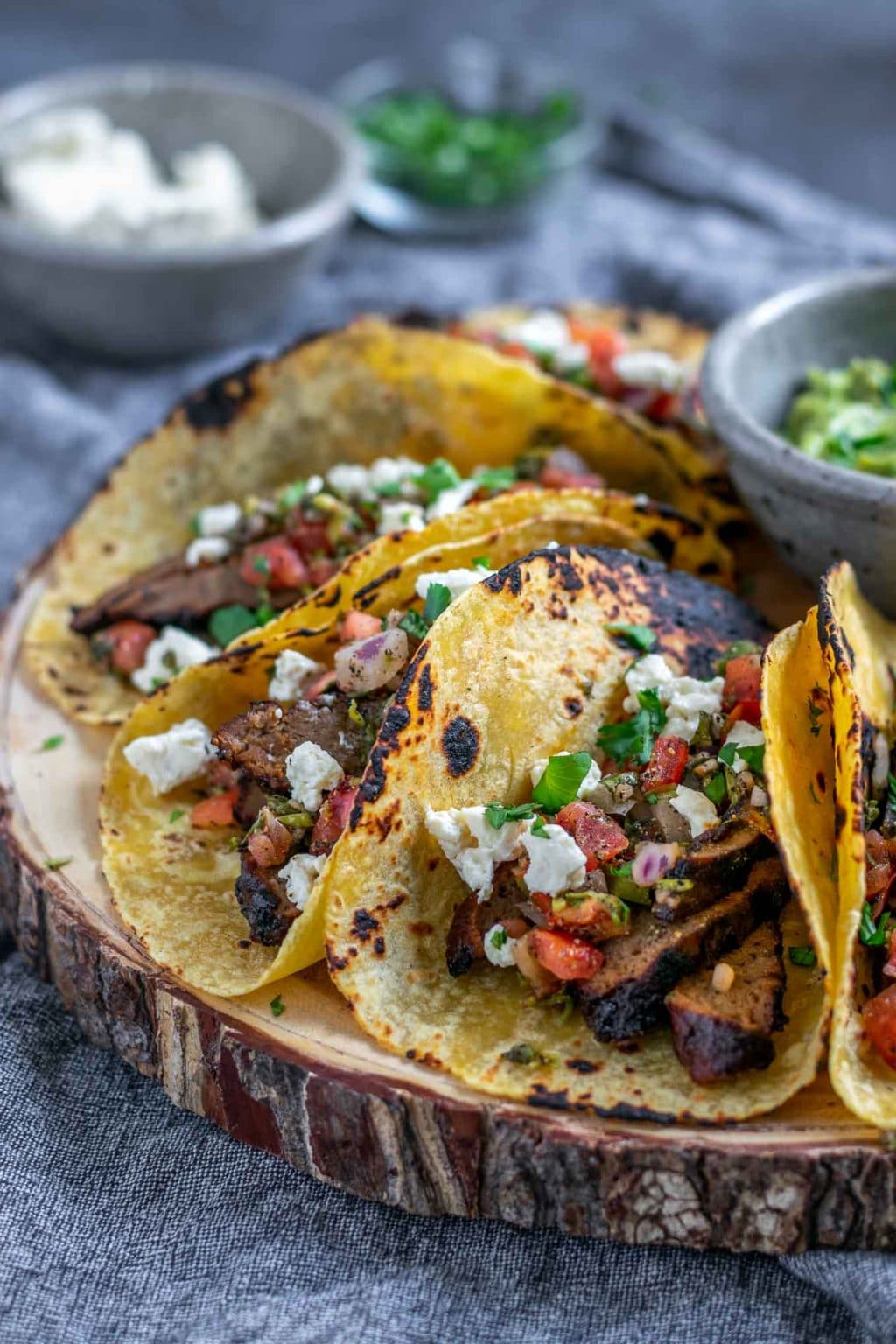 Vegan seitan carne asada tacos topped with pico de gallo and vegan queso fresco with guacamole and extra marinade, queso fresco, and cilantro to serve. 5 tacos assembled on a platter, side view.