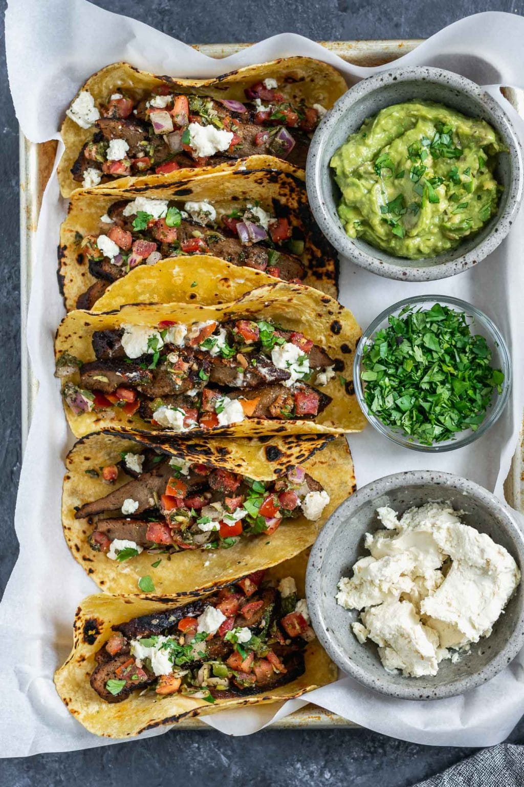 Vegan seitan carne asada tacos topped with pico de gallo and vegan queso fresco with guacamole, queso fresco, and cilantro to serve. 5 tacos assembled on a tray.