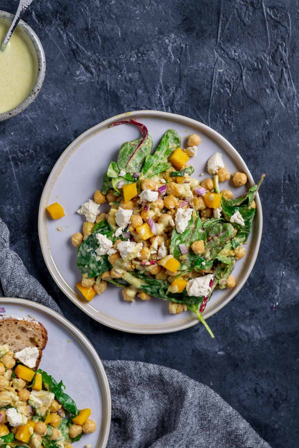 Curried chickpea and mango salad with vegan queso fresco. Extra dressing in a bowl in top corner and a second plate of the salad with a slice of toast