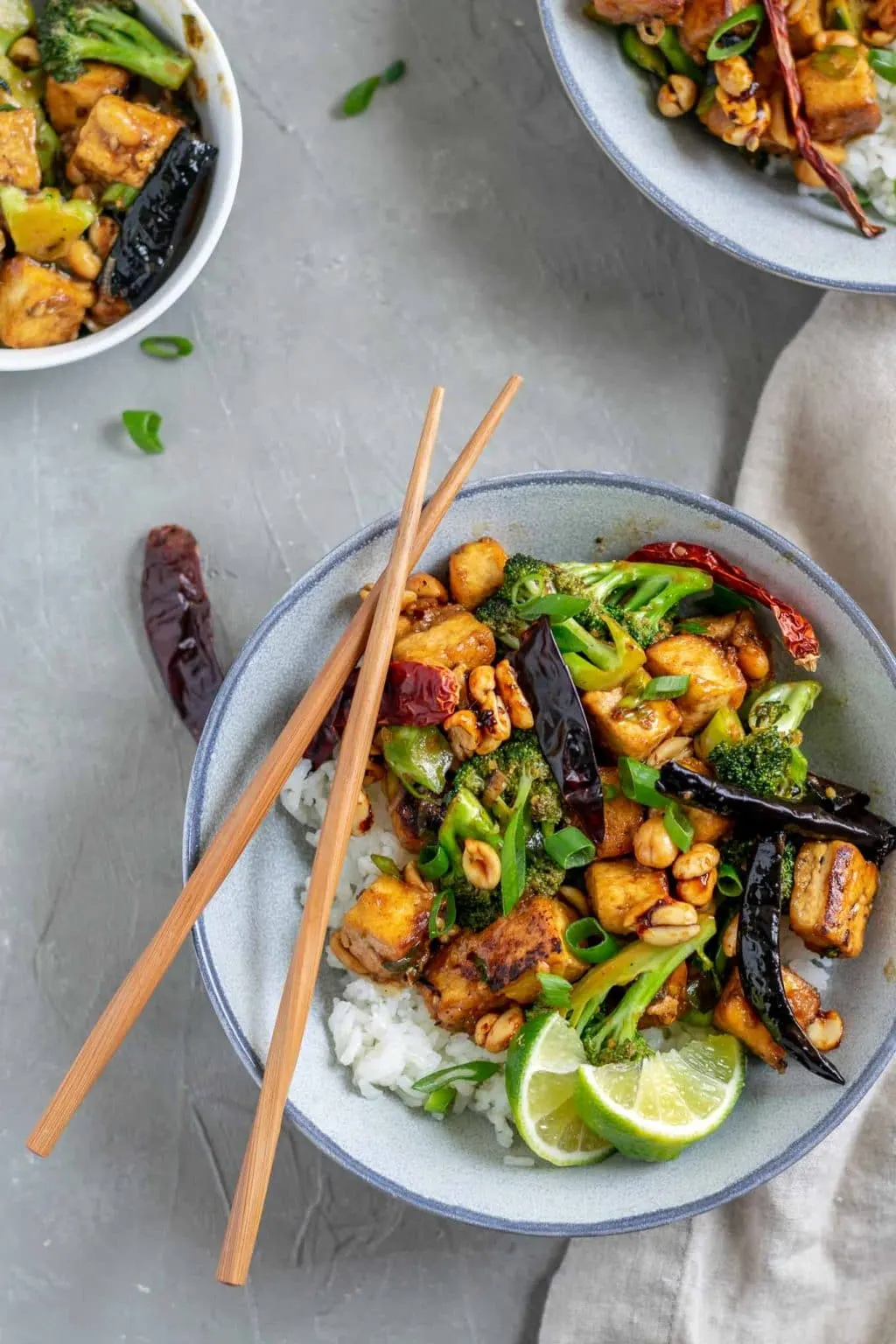 vegan kung pao tofu with broccoli and jasmine rice. Garnished with lime wedges and scallion greens and a pair of chopsticks is set over the bowl