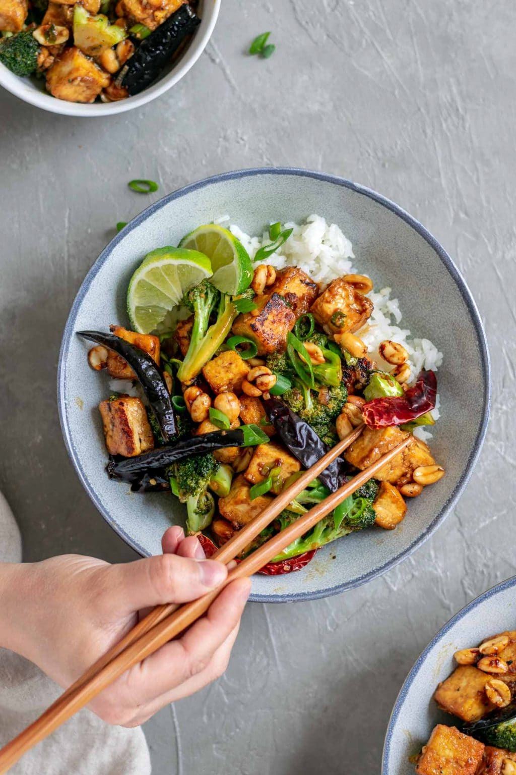 vegan kung pao tofu with broccoli and jasmine rice. Garnished with lime wedges and scallion greens. Chopsticks are being used to pick up a piece of tofu. 