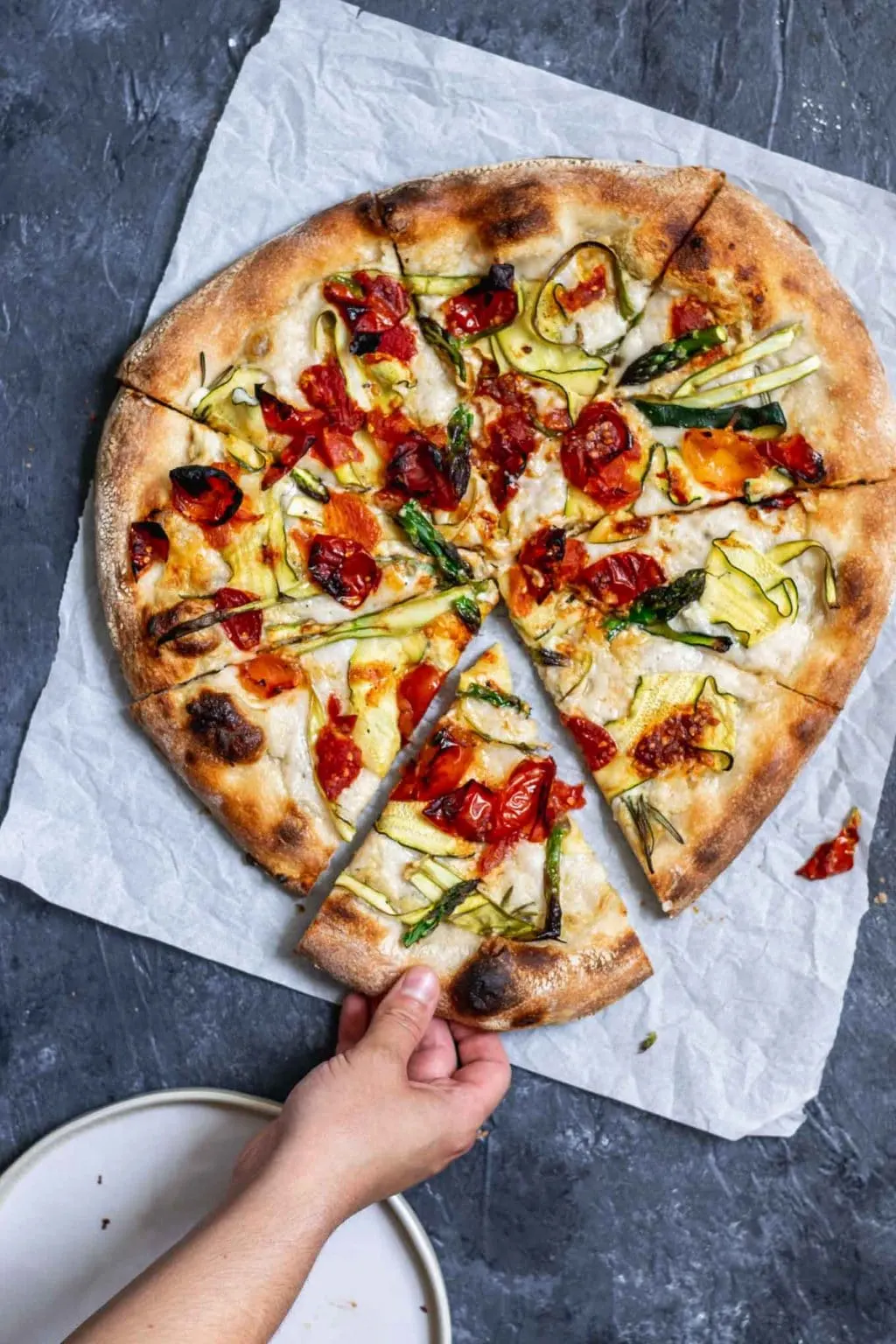 Grabbing a slice of vegan pizza primavera with zucchini, asparagus, and burst cherry tomatoes.