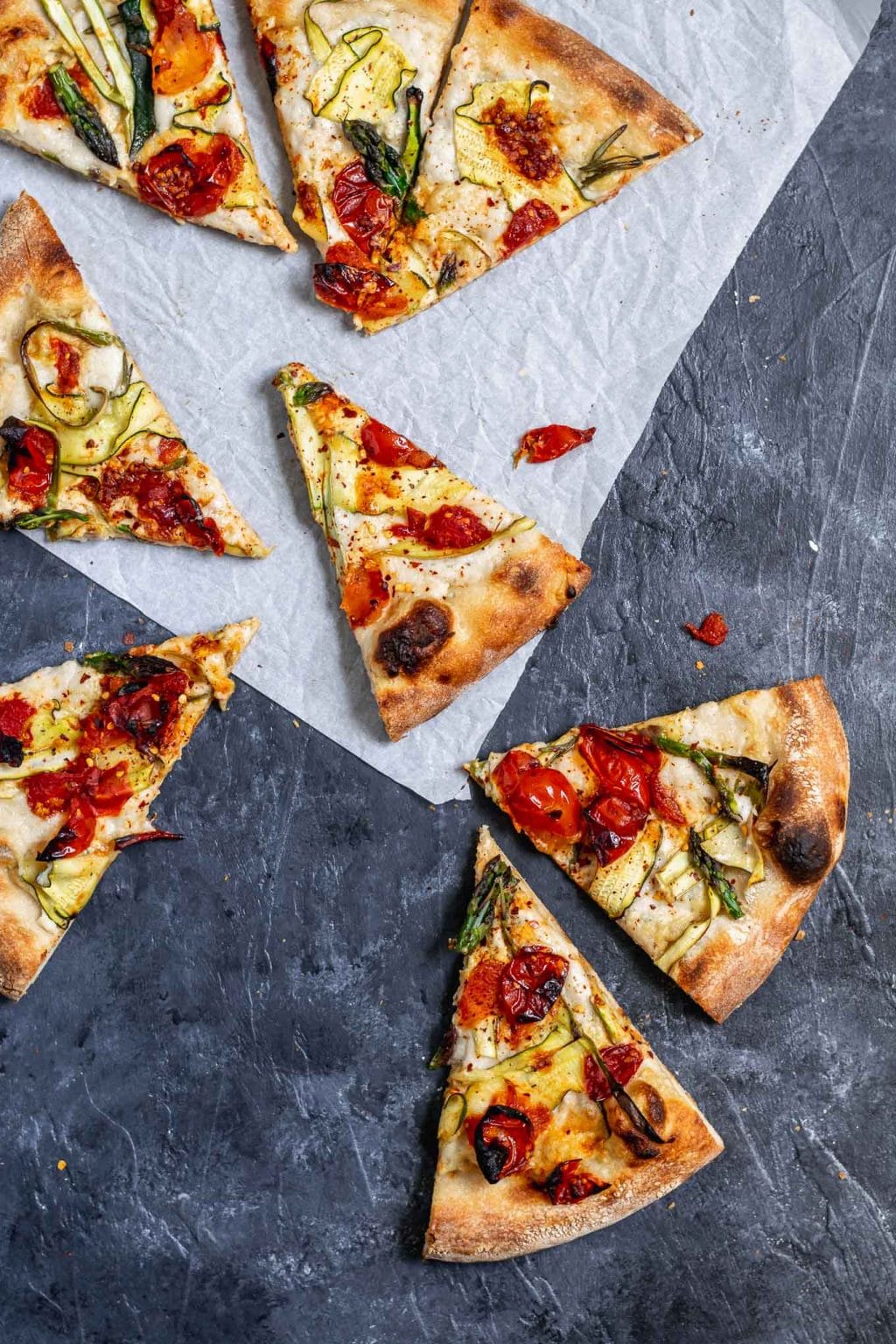 vegan pizza primavera with zucchini, asparagus, and burst cherry tomatoes. Overhead photo with pizza cut into slices arranged tumbling off a parchment paper.