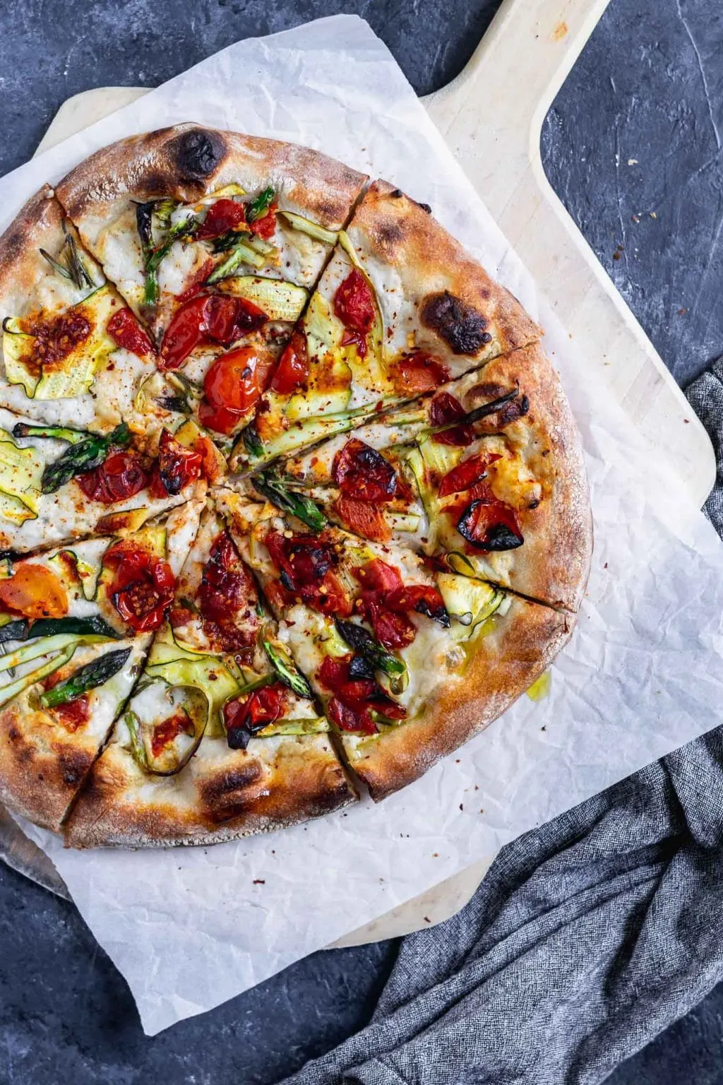 vegan pizza primavera with zucchini, asparagus, and burst cherry tomatoes. Overhead photo with pizza cut into slices on a parchment paper on top of a wooden pizza peel.