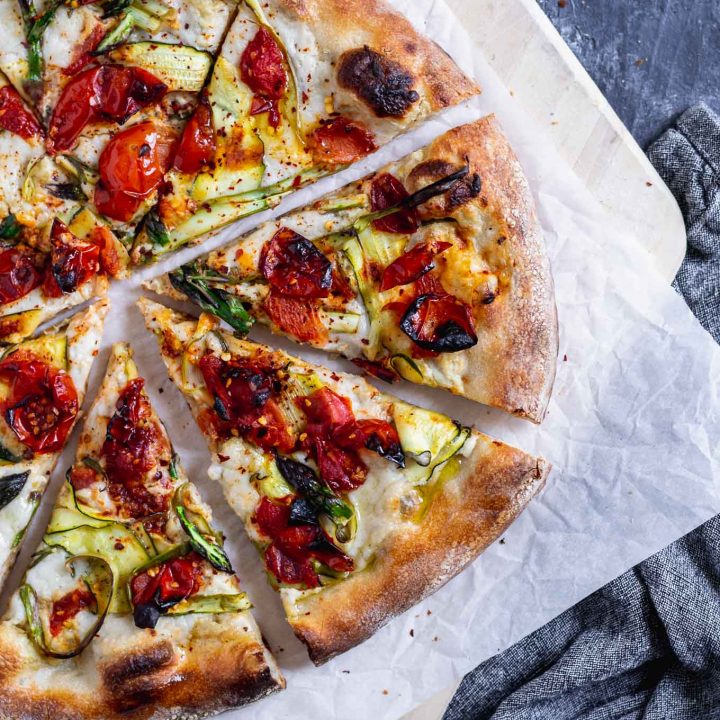 vegan pizza primavera with zucchini, asparagus, and burst cherry tomatoes. Overhead photo with pizza cut into slightly pulled apart slices on a parchment paper on top of a wooden pizza peel.