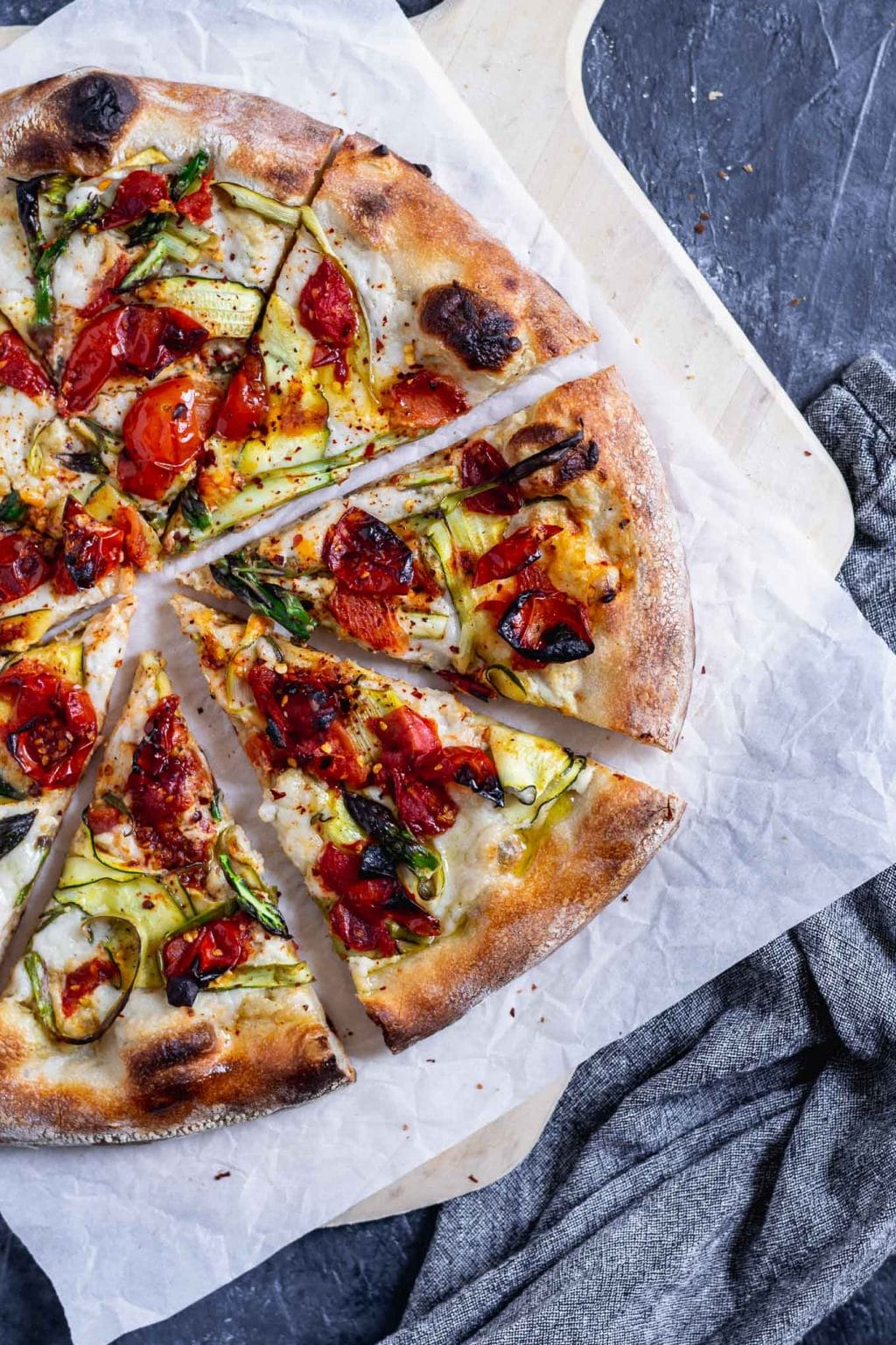 vegan pizza primavera with zucchini, asparagus, and burst cherry tomatoes. Overhead photo with pizza cut into slightly pulled apart slices on a parchment paper on top of a wooden pizza peel.