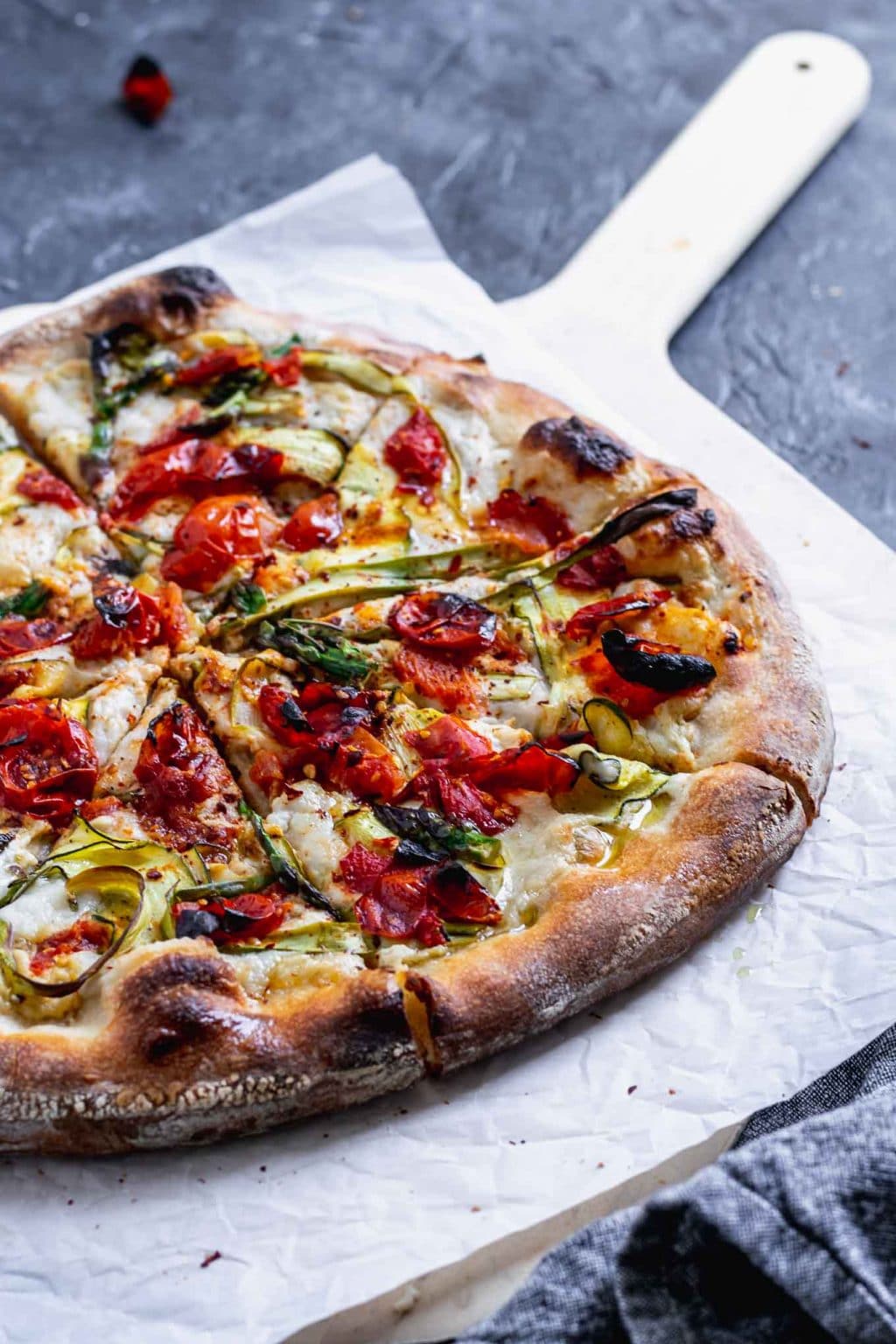 vegan pizza primavera with zucchini, asparagus, and burst cherry tomatoes. Angled view with pizza cut into slices on a parchment paper on top of a wooden pizza peel.