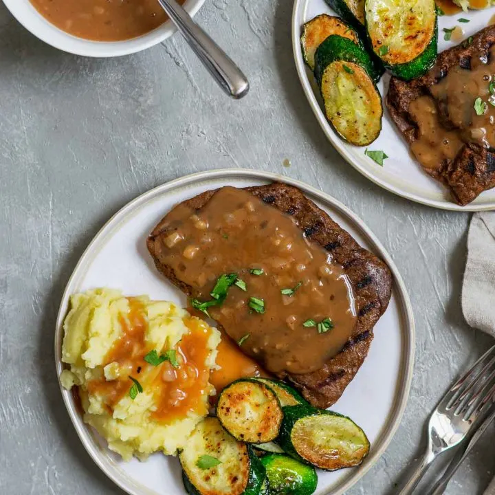 Grilled homemade seitan steaks with mashed potatoes and shallot gravy with sautéed zucchini, garnished with parsley, and extra gravy on the side