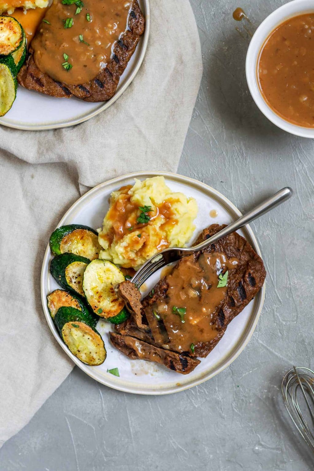 Bistecche di seitan alla griglia con purè di patate e salsa allo scalogno con un contorno di zucchine e una guarnizione di prezzemolo. La bistecca viene tagliata con un solo morso sulla forchetta e c'è del sugo extra servito a lato.