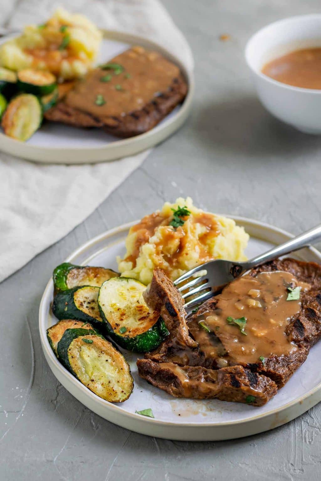 Grilled homemade seitan steaks with mashed potatoes and shallot gravy with a side of zucchini and a garnish of parsley. The steak is cut with one bite on a fork and there is extra gravy served on the side.