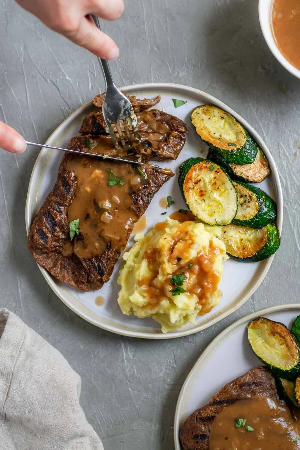 Grilled homemade seitan steaks with mashed potatoes and shallot gravy with a side of zucchini and a garnish of parsley. The steak is being cut with a knife and fork with hands in the photo and there is extra gravy served on the side.