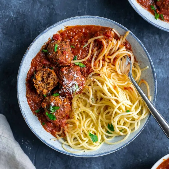 Spaghetti with roasted eggplant marinara and vegan lentil meatballs, one lentil meatball split open to show texture. Garnished with vegan parmesan and minced parsley. With fork twirling spaghetti in the bowl.