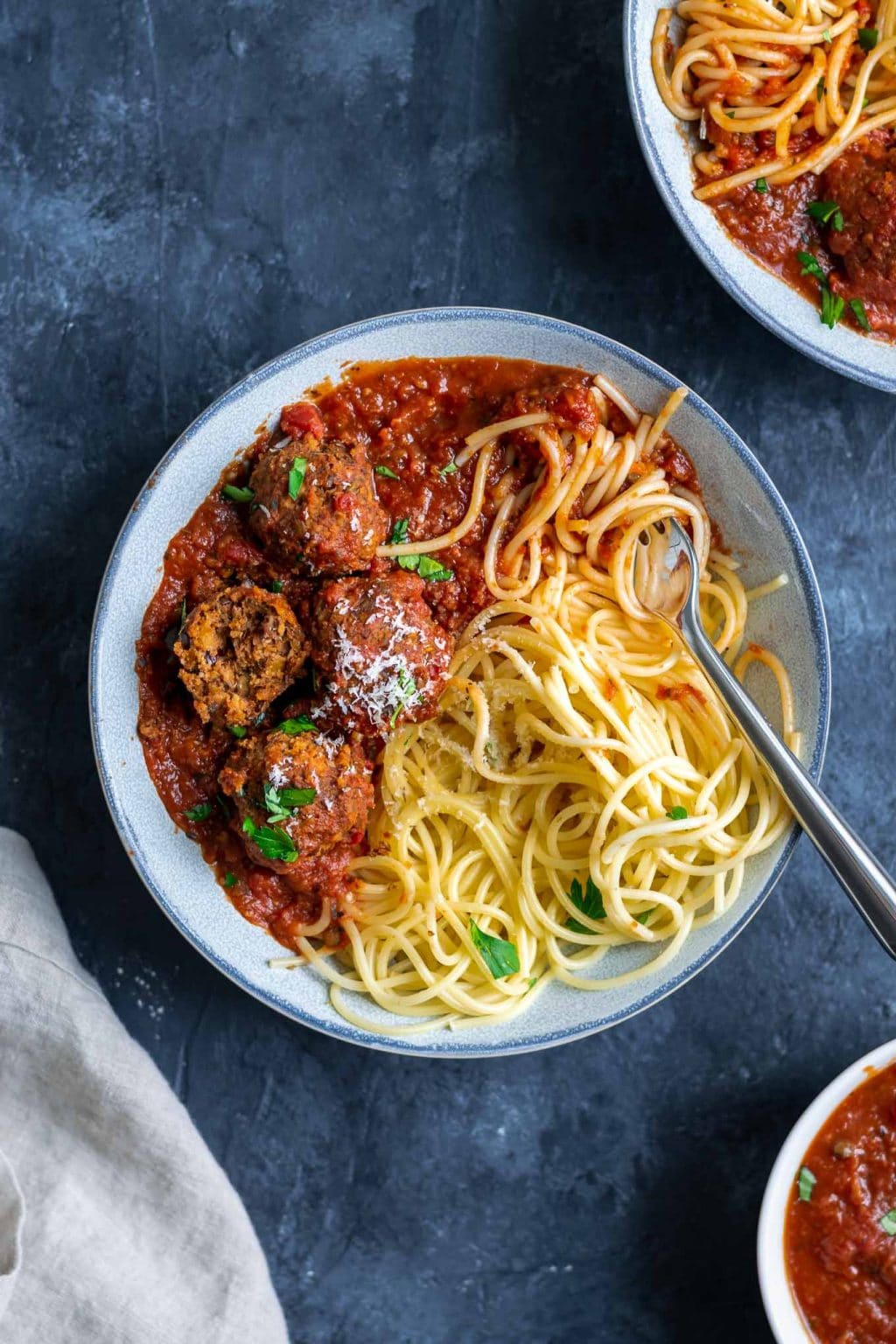 Spaghetti with roasted eggplant marinara and vegan lentil meatballs, one lentil meatball split open to show texture. Garnished with vegan parmesan and minced parsley. With fork twirling spaghetti in the bowl.