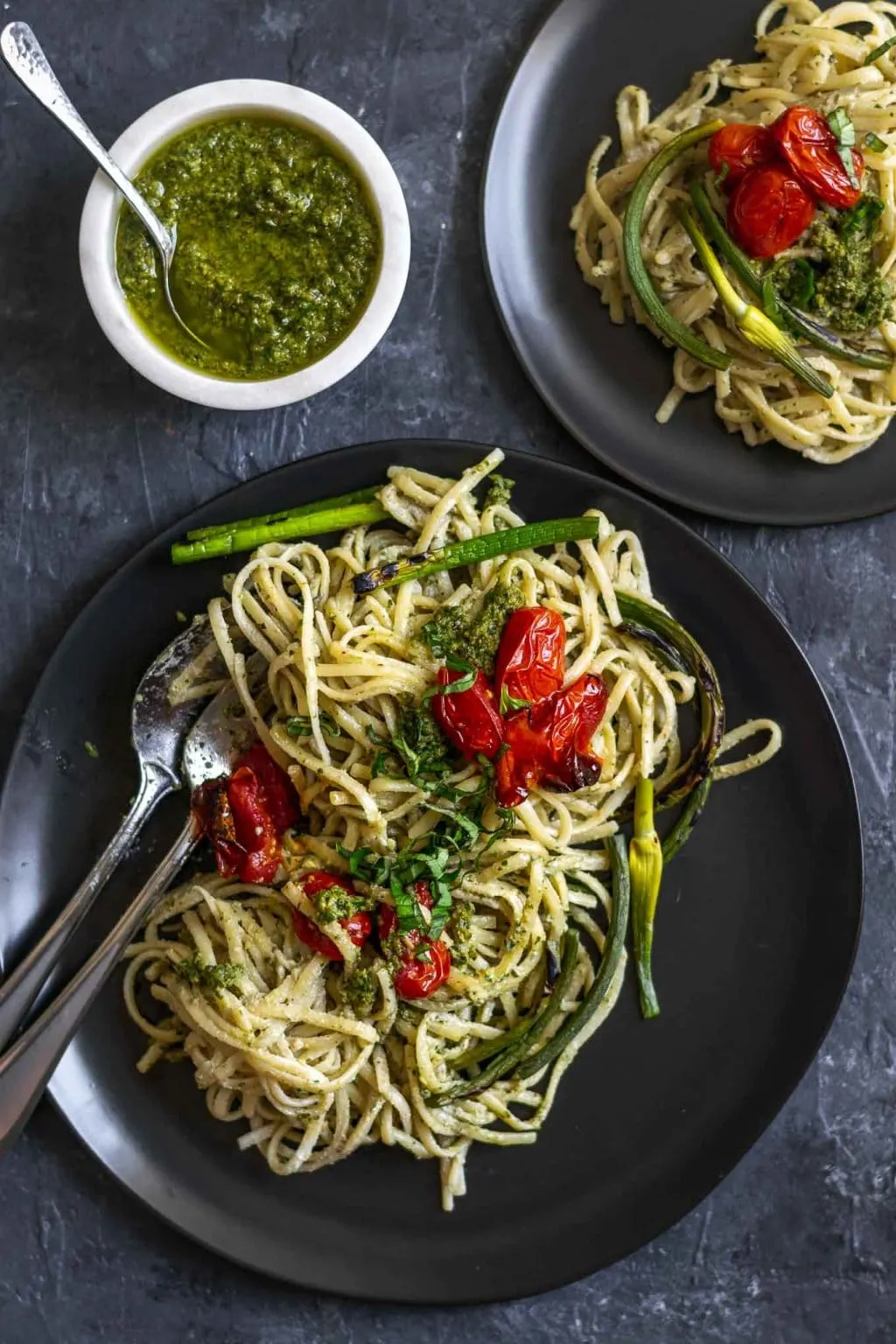vegan garlic pesto alfredo pasta with grilled garlic scapes and roasted cherry tomatoes served on two plates with extra garlic scape pesto