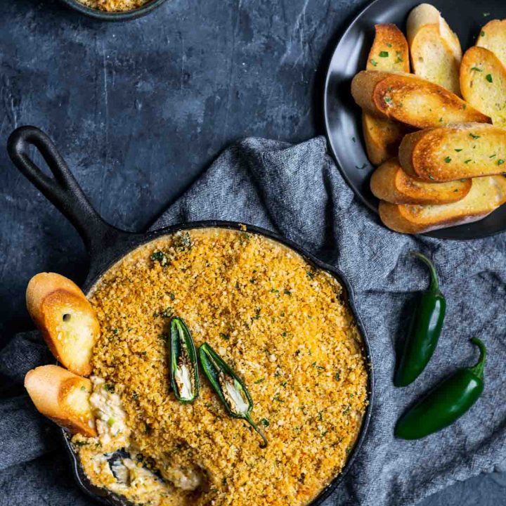 vegan jalapeño popper dip baked in a cast iron with a breadcrumb topping and crostini dipped into the pan with extra on the side