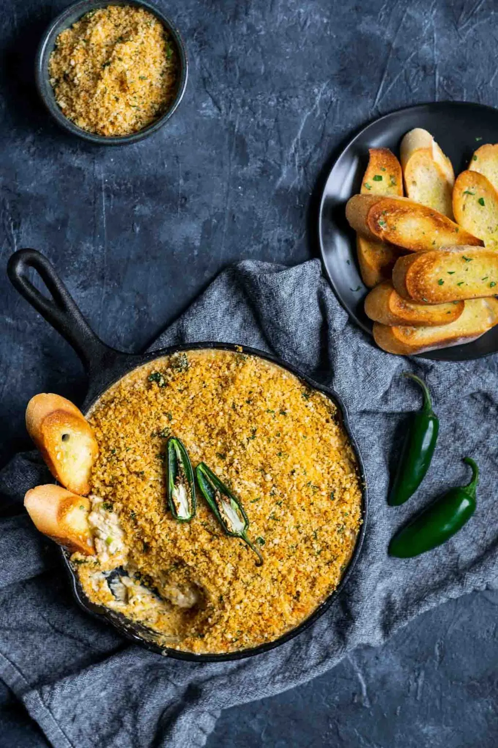 vegan jalapeño popper dip baked in a cast iron with a breadcrumb topping and crostini dipped into the pan with extra on the side