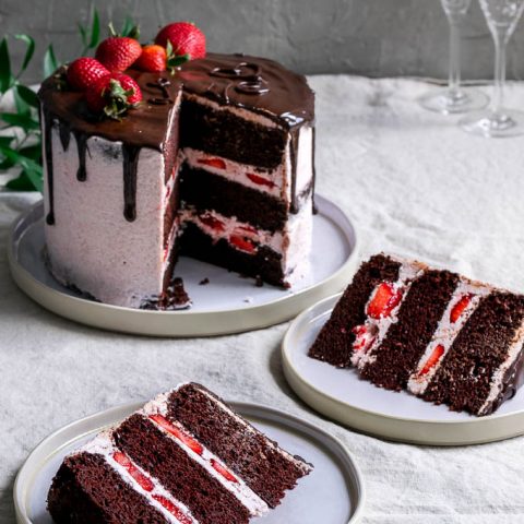 Two slices of the ultimate easy vegan chocolate cake with strawberry Italian meringue buttercream, a chocolate drip, and fresh strawberries to decorate