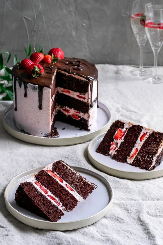 Two slices of the ultimate easy vegan chocolate cake with strawberry Italian meringue buttercream, a chocolate drip, and fresh strawberries to decorate