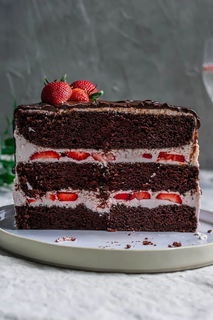 Cross section of the ultimate easy vegan chocolate cake with strawberry Italian meringue buttercream, a chocolate drip, and fresh strawberries to decorate