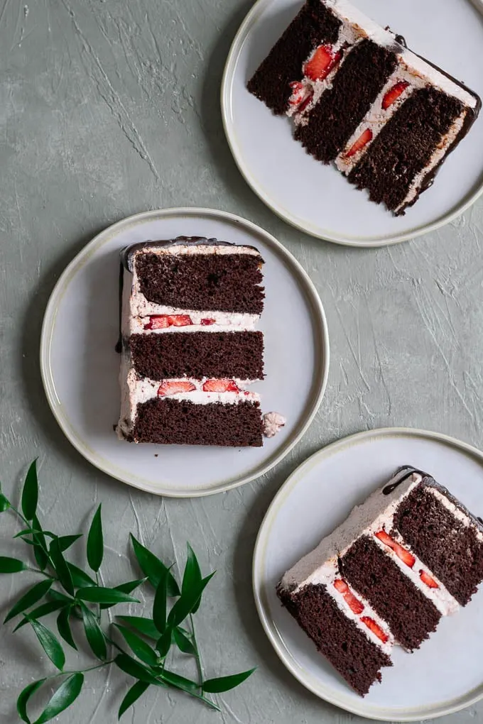 Three slices of the ultimate easy vegan chocolate cake with strawberry Italian meringue buttercream, a chocolate drip, and fresh strawberries to decorate