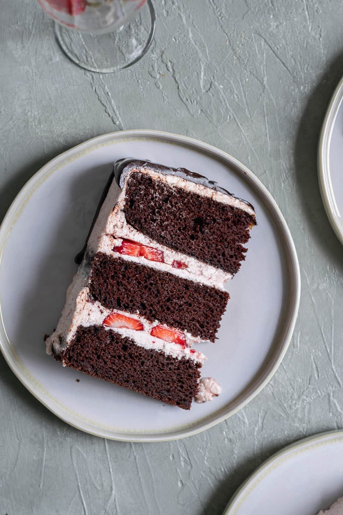 One slice of the ultimate easy vegan chocolate cake with strawberry Italian meringue buttercream, a chocolate drip, and fresh strawberries to decorate