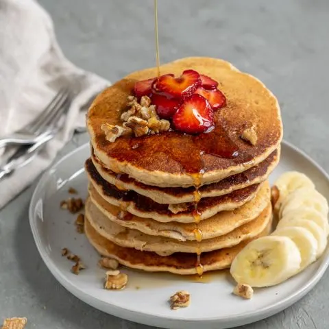 A stack of whole grain cornmeal vegan pancake mix, cooked and served with sliced strawberries, chopped walnuts, sliced banana, and a drizzle of maple syrup