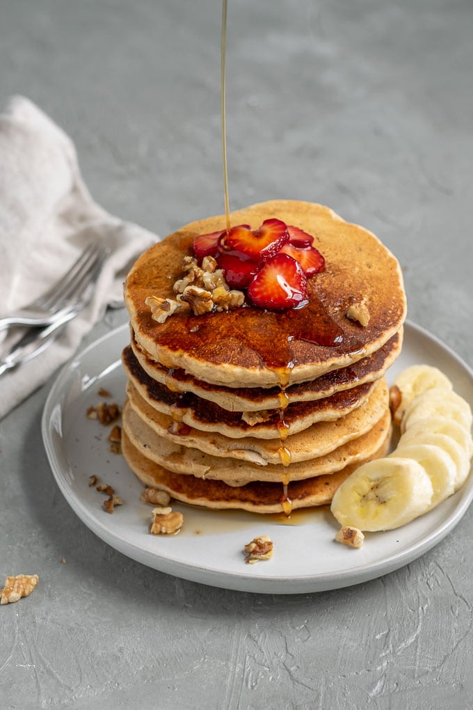 drizzling maple syrup over a stack of whole grain cornmeal vegan pancakes which are stacked 6 tall and served with sliced strawberries, chopped walnuts, and sliced banana