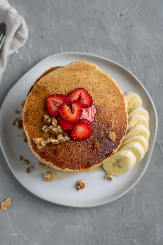 overhead view of a stack of make ahead whole grain cornmeal vegan pancakes topped with strawberry, additional walnuts and served with banana