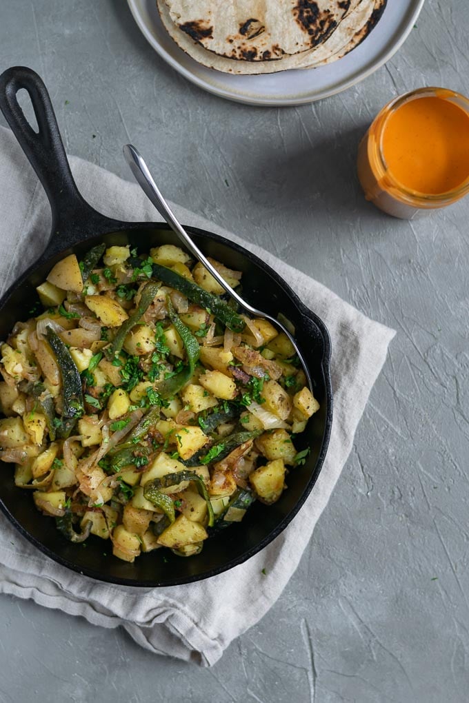 Paps rajas (potato and roasted poblano peppers) served in a cast iron skillet with corn tortillas and a side of creamy Mexican chipotle sauce