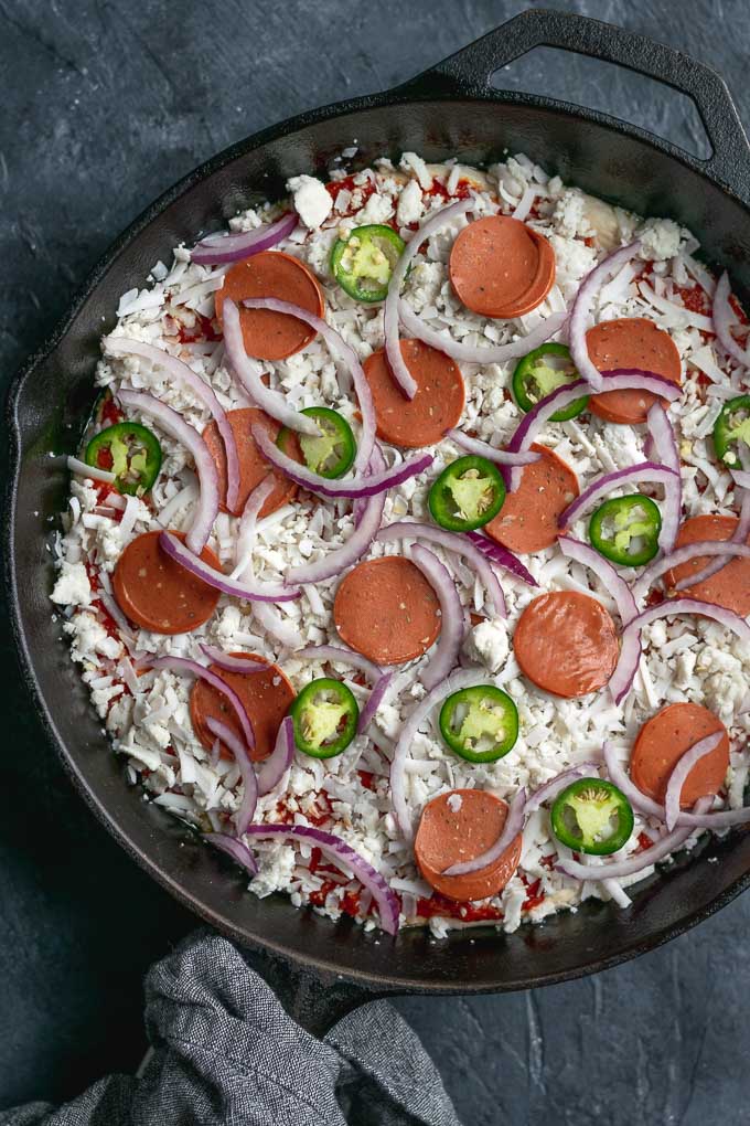 assembling vegan pepperoni pan pizza, topped with vegan pepperoni, vegan mozzarella, sliced jalapeños, sliced red onion