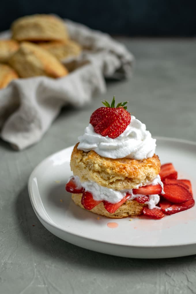 vegan strawberry shortcake with brandy soaked strawberries and vanilla bean whipped cream topped with a strawberry and extra shortbread biscuits in background