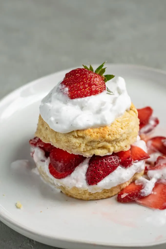 Close up of vegan strawberry shortcake with brandy soaked strawberries and vanilla bean whipped cream topped with a strawberry. A little messy with strawberries spilling out of the biscuit.