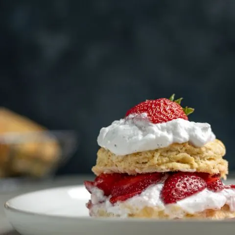 vegan strawberry shortcake with brandy soaked strawberries and vanilla bean whipped cream topped with a strawberry straight on close up shot. Extra biscuits in background.
