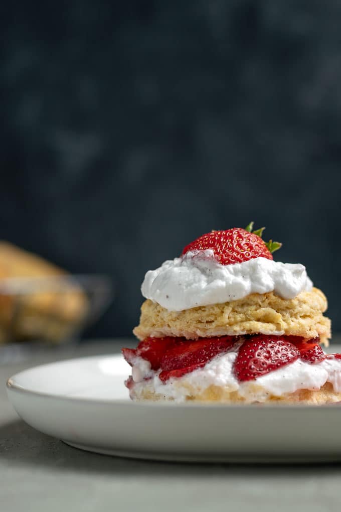 vegan strawberry shortcake with brandy soaked strawberries and vanilla bean whipped cream topped with a strawberry straight on close up shot. Extra biscuits in background.