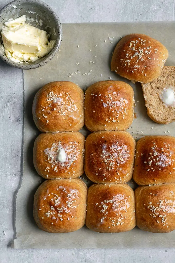 soft whole wheat dinner rolls on a parchment, one cut open with a pat of vegan butter on it