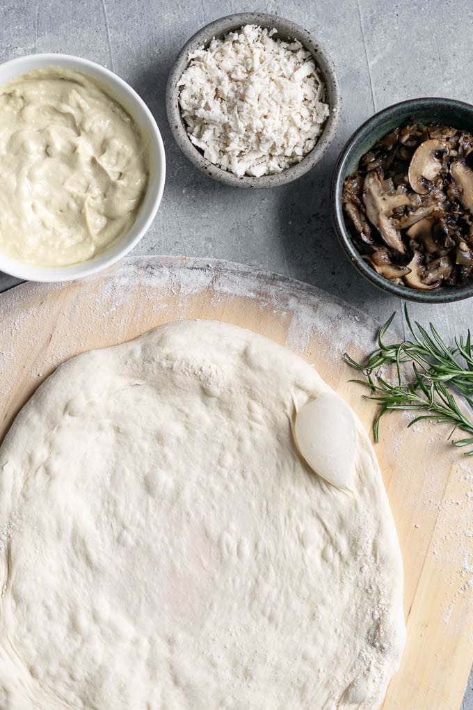 assembling the pizza with garlic white sauce, caramelized mushrooms, vegan mozzarella and fresh rosemary