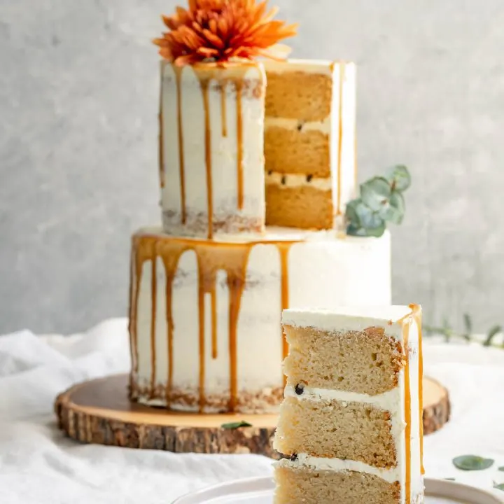 A 2-tier easy vegan vanilla cake decorated with flowers and a salted caramel drip with one slice cut out of it to reveal a 3 layer cake with chocolate chips in the buttercream filling