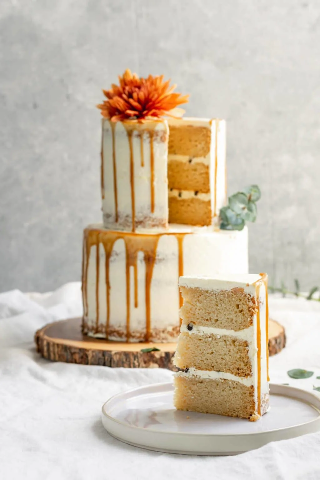 A 2-tier easy vegan vanilla cake decorated with flowers and a salted caramel drip with one slice cut out of it to reveal a 3 layer cake with chocolate chips in the buttercream filling